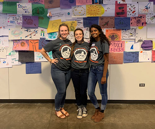  Elena Kamis, Angelina Boynton, and Shivani Ganesh, members of ISS's Educational Outreach Board
 at the fall 2019 Illinois Space Day hosted by ISS. In the background is some of the space artwork completed by young visitors to Illinois Space Day.